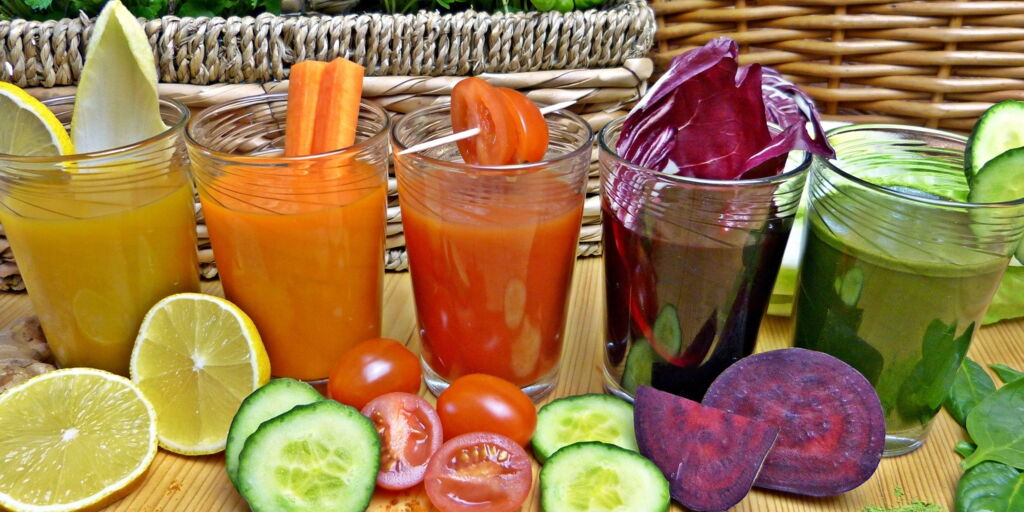 fünf Gläser mit frisch zubereitetem Gemüsesaft in unterschiedlichen Farben, davor liegen Stücke der Zutaten, z.B. Zitrone, Gurke, Tomate, rote Beete