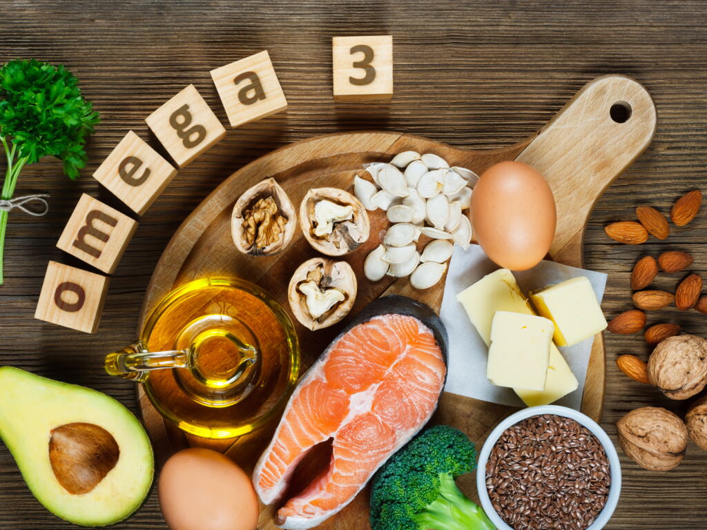 Omega 3-haltige Lebensmittel tierisch und pflanzlich (Nüsse, Leinsaat, Mandeln, Avocado, Ei, Butter, Lachs, Öl) auf Holzplatte angerichtet