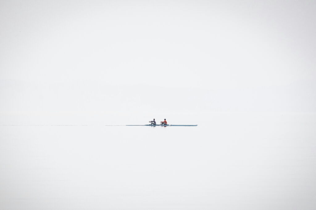 weit entfernt zwei Ruderer in schmalem Boot auf spiegelglattem, ruhigen See, diffuses Licht