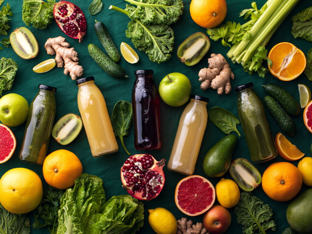 Zum Saftpressen geeignetes frisches Obst und Gemüse, mit 5 Flaschen gepresstem Saft auf dunklem Untergrund (Grapefruit, Apfel, Zitrone, Kiwi, Gurke, Salat, Stangensellerie,...)