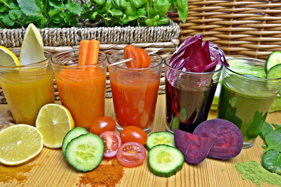 fünf Gläser mit frisch zubereitetem Gemüsesaft in unterschiedlichen Farben, davor liegen Stücke der Zutaten, z.B. Zitrone, Gurke, Tomate, rote Beete