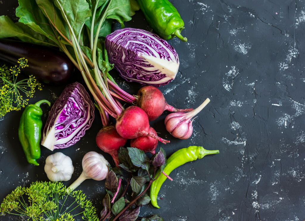gemischtes rohes Gemüse und Kräuter auf dunklem Hintergrund (rote Beete mit Blättern, Rotkohl aufgeschnitten, Knoblauknollen, Fencheldolden,...)