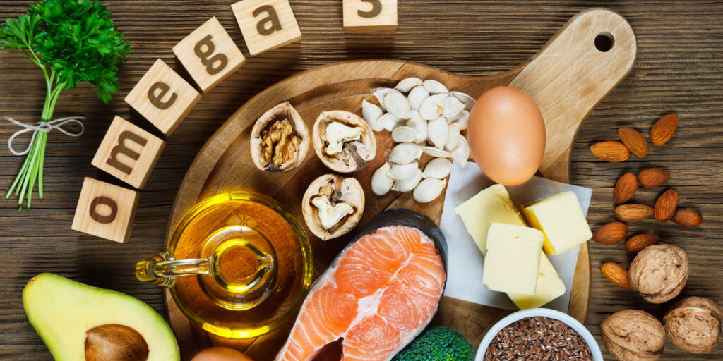 Omega 3-haltige Lebensmittel tierisch und pflanzlich (Nüsse, Leinsaat, Mandeln, Avocado, Ei, Butter, Lachs, Öl) auf Holzplatte angerichtet