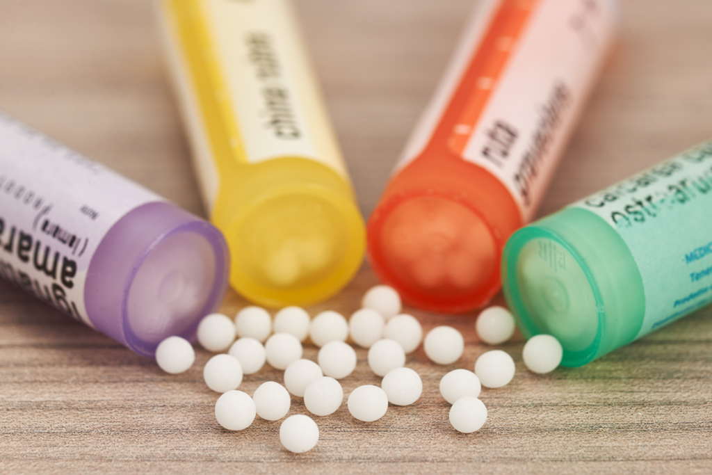 vier farbige Röhrchen für homöopathische Mittel auf Tischplatte. Mehrere Globuli weiße Kügelchen im Vordergrund