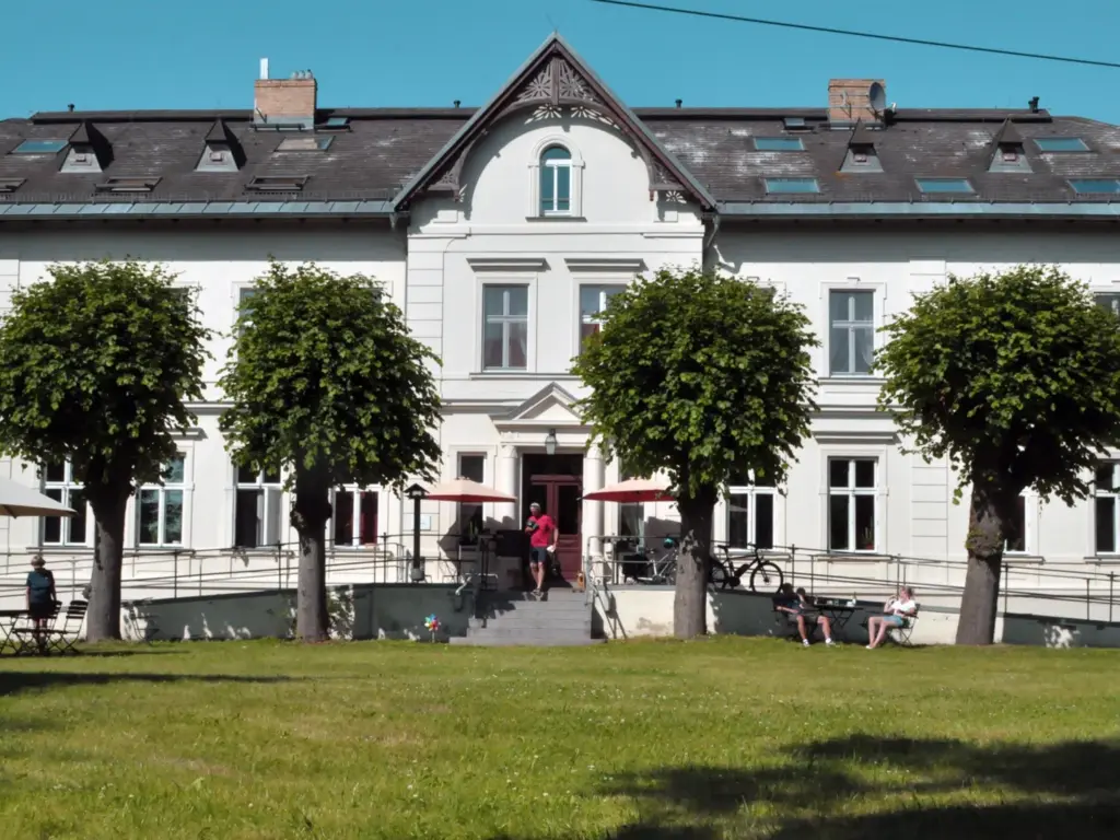 sommerliche Vorderansicht historisches Gutshaus, zweistöckig, kleine Freitreppe in der Mitte, davor Wiese und kugelig geschnittene Lindenbäume