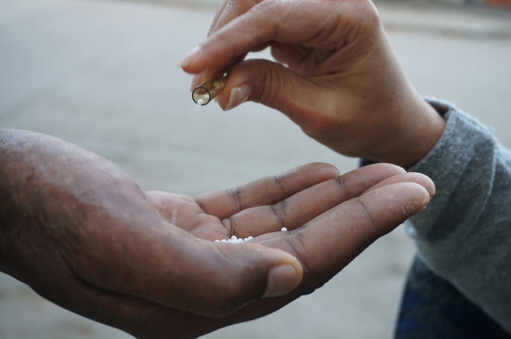 Eine Person gibt homöopathische Globuli aus einem Röhrchen in die offene Hand einer anderen Person.