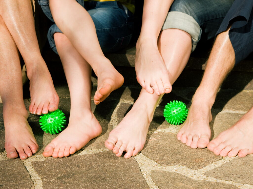 Detailansicht Beine und Füße vier auf Treppenstufen sitzende Menschen mit hochgerollten Hosenbeinen und nackten Füßen mit zwei grünen Igel-Gymnastikbällen