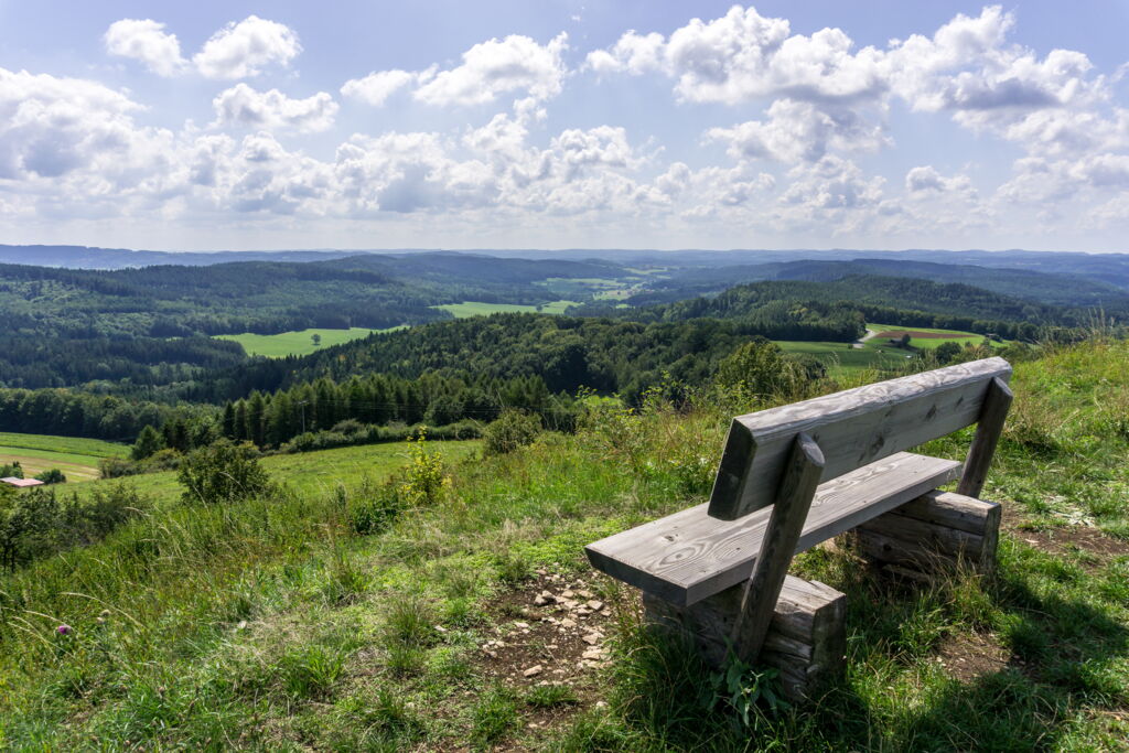 rustikale Holzbank auf Anhöhe mit weitem Blick in hügelige Landschaft, Wald, Wiesen, Sonnenschein, Himmel mit kleinen Schönwetterwolken rustikale Holzbank auf Anhöhe mit weitem Blick in hügelige Landschaft, Wald, Wiesen, Sonnenschein, Himmel mit kleinen Schönwetterwolken