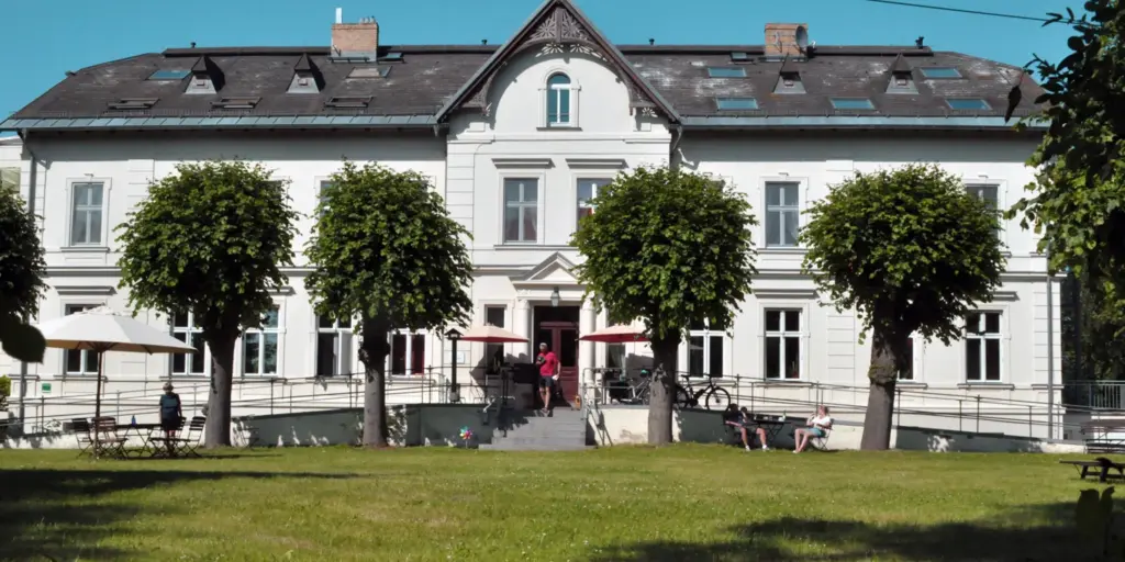 sommerliche Vorderansicht historisches Gutshaus, zweistöckig, kleine Freitreppe in der Mitte, davor Wiese und kugelig geschnittene Lindenbäume