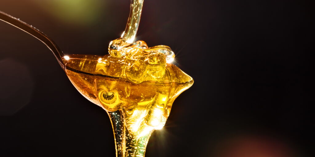 goldener Honig fliesst in einen Löffel, sammelt sich dort, fliesst weiter vor dunklem Hintergrund