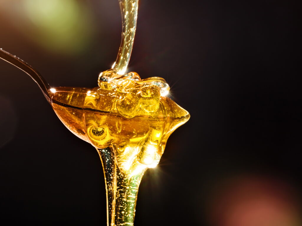 goldener Honig fliesst in einen Löffel, sammelt sich dort, fliesst weiter vor dunklem Hintergrund