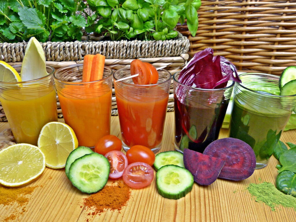 fünf Gläser mit frisch zubereitetem Gemüsesaft in unterschiedlichen Farben, davor liegen Stücke der Zutaten, z.B. Zitrone, Gurke, Tomate, rote Beete