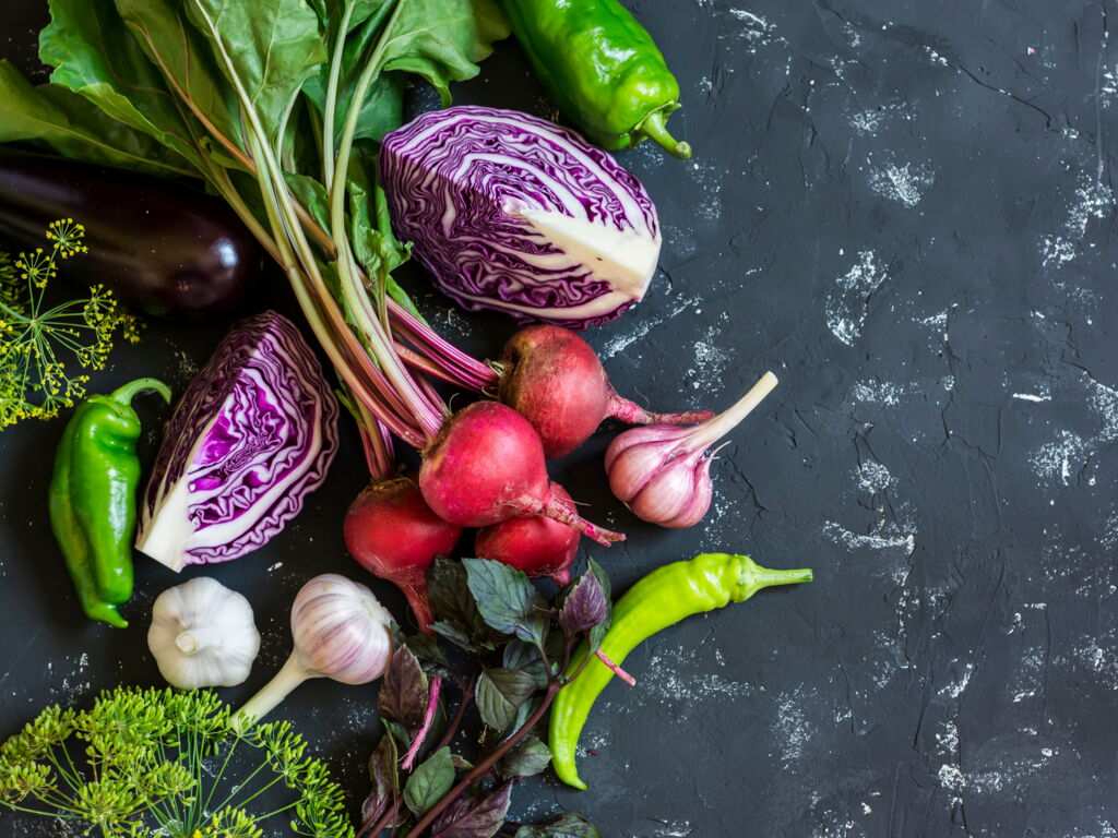 gemischtes rohes Gemüse und Kräuter auf dunklem Hintergrund (rote Beete mit Blättern, Rotkohl aufgeschnitten, Knoblauknollen, Fencheldolden,...)